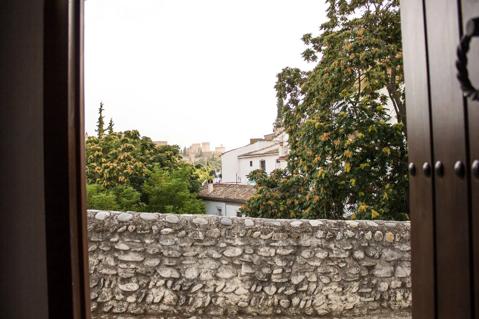 Puerta de la casa abierta a un muro de piedra, con casas del barrio y la Alhambra al fondo entre los árboles